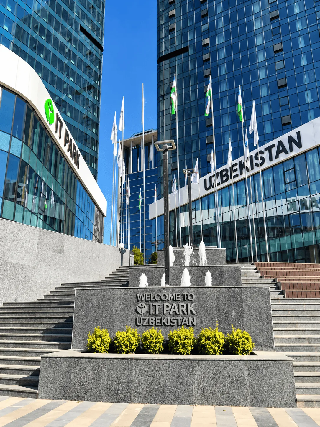 Вход в IT Park Uzbekistan: приветственная надпись, фонтан и флаги перед стеклянными башнями